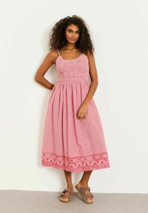 Frau mit lockigem Haar, die ein rot-weiß kariertes Midikleid mit Spaghettiträgern und braune Sandalen trägt, steht vor einer weißen Wand.