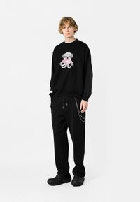 Sudadera negra con un gráfico de oso de peluche en rosa y gris, combinada con pantalones negros y calzado texturizado negro.