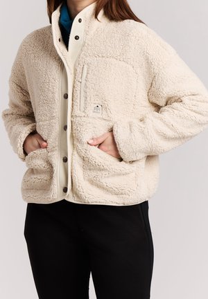 Femme portant une veste en sherpa polaire crème avec boutons devant et poches, mains dans les poches, pantalon noir visible.