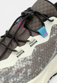 Καφέ αθλητικό παπούτσι με υφή και λευκή σόλα, με μαύρα κορδόνια, ροζ και μπλε λεπτομέρειες και σχεδιασμένη επάνω επιφάνεια.