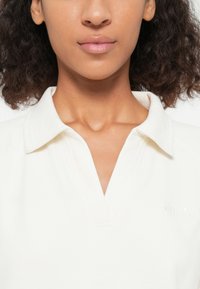 Sudadera de cuello blanco con un diseño de escote en V, que presenta un sutil logo en relieve en el lado izquierdo y una tela texturizada.