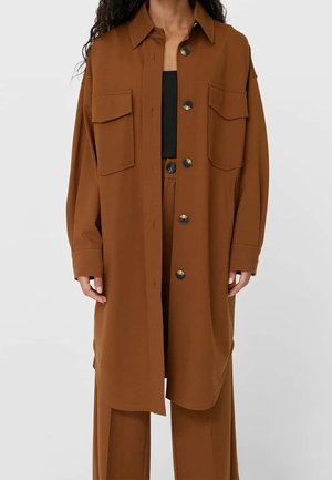 Femme portant une longue veste-chemise marron boutonnée avec deux poches poitrine sur un pantalon large assorti et un haut noir.