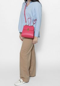 Roze leren handtas met een gestructureerd ontwerp, gouden hardware en een afneembare schouderriem. Draagt bij een lichtblauw shirt en gestreepte broek.