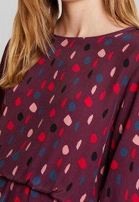 Fialová tkanina s abstraktními tvary v červené, růžové, modré a černé barvě. Hladký povrch s volným střihem.