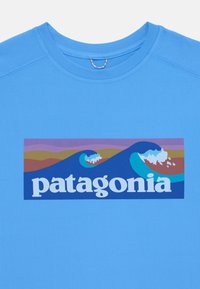 Gaismas zila T-krekls ar stilizētu viļņu un kalnu grafiku, kurā burtu ģimenē ar lieliem, baltajiem burtiem ir vārds "patagonia".