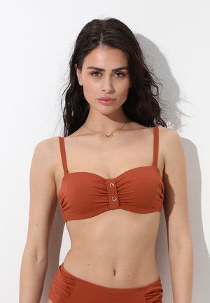 Donna con capelli lunghi e scuri che indossa un top bikini a fascia color ruggine con spalline sottili, in piedi su sfondo bianco.