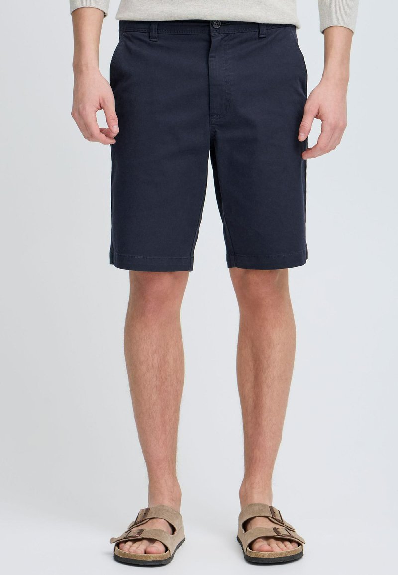 Shorts en coton bleu marine à coupe droite, avec poches avant et fermeture à bouton. Portés avec des sandales en suède marron avec boucles.