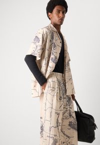 Beige outfit with map print; short-sleeve shirt and wide-leg pants. Black long-sleeve shirt underneath. Model holds a black bag.