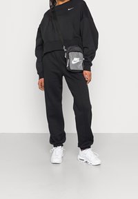 Person som bär en svart Nike sweatshirt och byxor, vita Nike sneakers och en liten svart-grå Nike crossbody-väska.