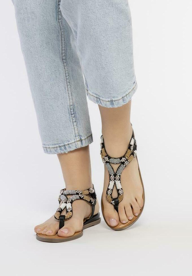Sandalias de flip-flop con cuentas negras y marrones, con un diseño de trenzado intrincado y suelas planas, llevadas con pantalones de mezclilla cortos claros.