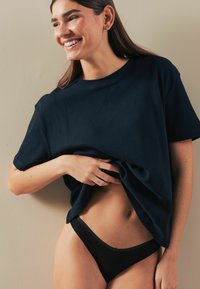 Tamno plava rebrasta majica s kratkim rukavima, širokog kroja, nošena preko crnih bikinija. Glatka tekstura, suptilni logotip branda na majici.