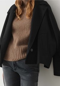 Schwarze Jacke mit strukturierter Silhouette, matter Textur und übergroßen Revers, kombiniert mit einem braunen, gerippten Strickpullover und grauer Jeans.
