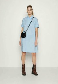 Mulher em vestido de manga curta azul claro com bolsa transversal preta e botas de salto alto castanhas até o tornozelo, em pé contra um fundo branco.