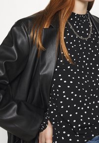 Blazer in pelle nera abbinato a una blusa nera a pois. La blusa presenta un colletto arricciato e motivi circolari bianchi. Collana in catena argentata.