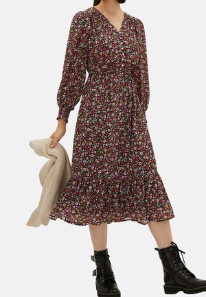 Frau trägt ein midilanges, langärmeliges Blumenkleid mit Rüschensaum, hält einen beigen Strickpullover und schwarze Schnürstiefeletten.