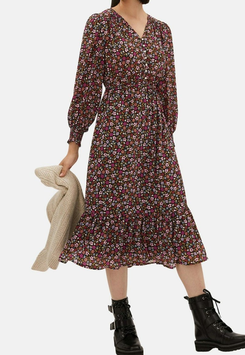Femme portant une robe mi-longue à manches longues à fleurs avec un ourlet volanté, tenant un pull beige en maille et des bottines noires à lacets.
