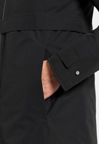 Hand inserted into side pocket of black jacket with buttoned cuff and visible zipper seam.
