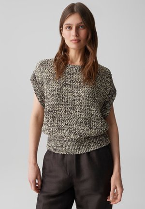 Jeune femme aux longs cheveux bruns portant un haut court en maille noir et blanc et un pantalon foncé, debout devant un fond gris uni.