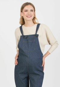 Dżinsowe ogrodniki z regulowanymi ramiączkami, kieszenią z przodu i kontrastującym przeszyciem, noszone na jasnobeżowym, prążkowanym, długim rękawie.