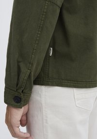 Olive green jacket with a buttoned cuff, featuring a visible logo tag at the hem. The fabric appears textured with a casual fit.
