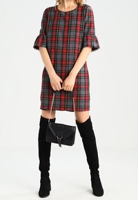 Robe à carreaux avec de courtes manches bouffantes en rouge, noir et jaune. Portée avec des bottes noires au-dessus du genou et un petit sac à main noir avec une chaîne.
