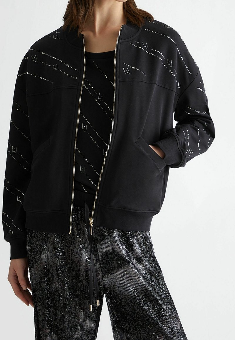 Giacca bomber nera con zip, caratterizzata da una texture liscia, un motivo diagonale di strass e tasche laterali, abbinata a pantaloni lucidi decorati con paillettes.
