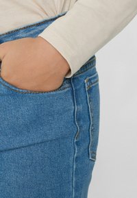 Džínové kalhoty s modrým nádechem, vybavené přední kapsou a kontrastním oranžovým stehem. Tkanina má měkkou texturu.