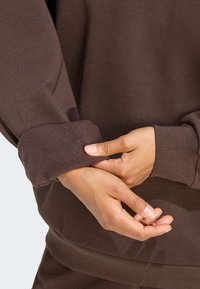 Mani che aggiustano il polsino di una felpa a maniche lunghe di colore marrone, foderata in fleece, mostrando la morbida texture del tessuto interno.
