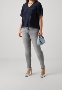 Marineblauwe geplooide blouse met V-hals, gecombineerd met grijze skinny jeans. Houdt een lichtblauwe handtas vast en draagt metalen zilveren puntige hakken.