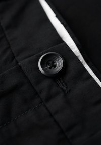 Botón negro cosido en tela negra con costura visible y un detalle de franja blanca a lo largo de la costura, botón grabado con "Superdry Studios."