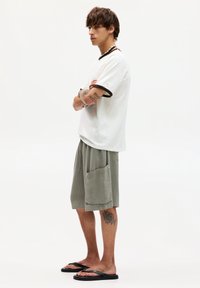 Hvid T-shirt med brun kant, grå teksturerede shorts med en sidelomme og sorte flip-flops. Tatoveringer synlige på armen og benet.