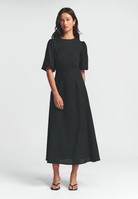 Černé maxi šaty s volánovými rukávy, upnutým živůtkem a rozšířenou sukní. Hladká tkanina s minimálními detaily, doplněné černými sandály.