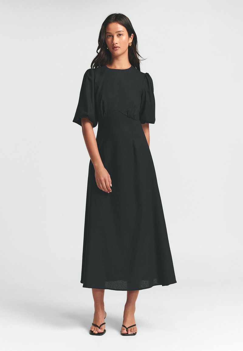 Schwarzes Maxikleid mit Puffärmeln, einem taillierten Oberteil und einem ausgestellten Rock. Glattes Material mit minimalen Details, kombiniert mit schwarzen Sandalen.