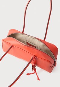 Prostokątna torebka z koralowej skóry, z długimi cienkimi paskami, srebrnym suwakiem, beżowym materiałowym wnętrzem oraz dekoracyjnym węzłem z koralowego sznura z klamrą.