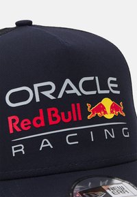 Mornarska kapa s izvezenim logotipima "Oracle" i "Red Bull Racing" u srebrnoj i crvenoj boji. Sadrži dva emblema crvene bika iznad žutog kruga.