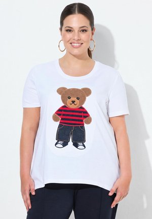Lachende vrouw met donker haar draagt grote oorringen, een wit T-shirt met een teddybeer in een rood gestreept shirt, spijkerbroek en sneakers.
