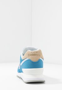 Sneaker en textile bleu avec talon en suède beige, accents blancs et un logo distinctif. Présente une semelle en caoutchouc et un col rembourré pour le confort.