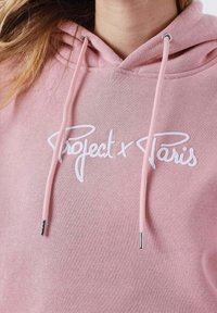 Sudadera con capucha rosa con texto bordado en blanco "Project x Paris". Cuenta con cordones con puntas metálicas y una tela suave y texturizada.