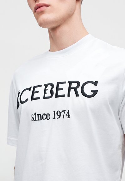 Άνδρας που φοράει λευκό t-shirt με μαύρο κεντημένο κείμενο "ICEBERG since 1974" στο στήθος σε απλή ανοιχτή περιοχή.
