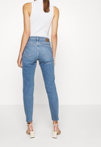Jeans ajustados de color azul claro con cintura alta, que presentan dos bolsillos traseros y una parche de cuero marrón en la cinturilla. El dobladillo está ligeramente deshilachado.