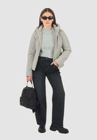 Giacca con cappuccio grigia chiara sopra un maglione grigio, abbinata a jeans neri a vita alta e stivali neri. Gli accessori includono uno zainetto nero piccolo.