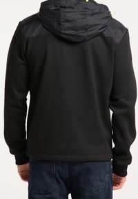 Czarna kurtka z kapturem i pikowanymi panelami na ramionach noszona przez osobę w niebieskich dżinsach, pokazana z tyłu na jednolitym tle.