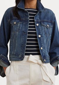 Denim jacket in dark blue with classic cut, fold-down collar, front pockets, and silver buttons. Worn over a navy and white striped top.