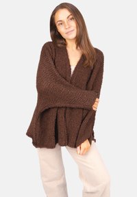 Femme aux longs cheveux bruns portant un cardigan sombre et texturé, ample, et un pantalon beige clair, se tenant devant un fond uni.
