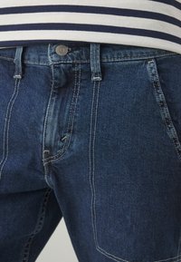 Jean en denim bleu foncé avec surpiqûres blanches contrastantes et un bouton en métal, porté avec une chemise rayée bleu marine et blanche.