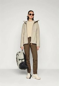 Beige vízálló kabát kapucnival, kötött bézs pulóver, barna nadrág és világos cipők. Szürke és fekete hátizsákot hord.