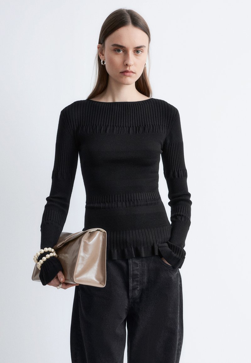 Kvinde iført en sort, tekstureret langærmet top og mørke bukser, der holder en stor beige clutch med perlearmbånd om håndleddet.