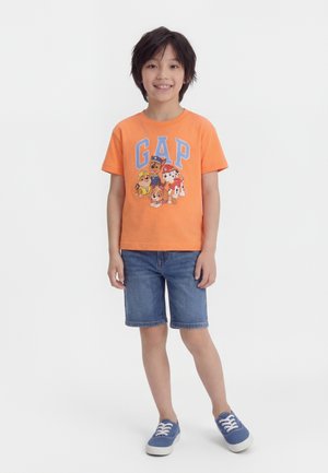 Χαμογελαστό παιδί που στέκεται με χαλαρά τα χέρια, φορώντας πορτοκαλί μπλουζάκι με το λογότυπο του Paw Patrol, τζιν σορτσάκι και μπλε αθλητικά παπούτσια σε λευκό φόντο.