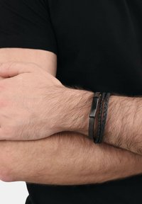 Braccia maschili incrociate, indossando una maglietta nera, con un braccialetto in pelle nera e due braccialetti intrecciati neri sul polso.