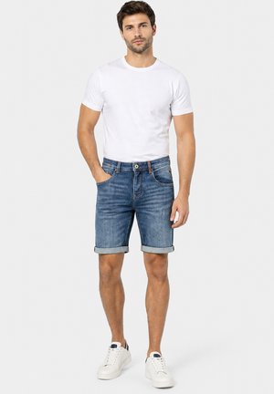Mann steht mit den Händen in den Taschen, trägt ein schlichtes weißes T-Shirt, hochgekrempelte blaue Jeansshorts und weiße Turnschuhe vor hellem Hintergrund.